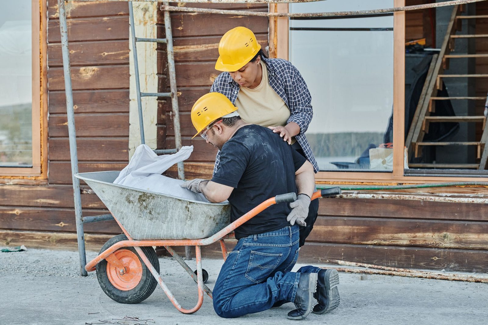 Un trabajador de la construcción se arrodilla junto a una carretilla en una obra mientras un compañero le ayuda.