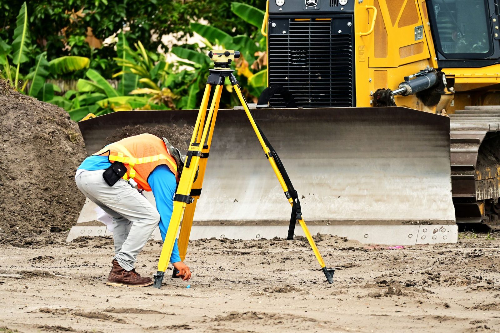 A construction worker uses a total station surveying instrument beside a bulldozer on a building site.