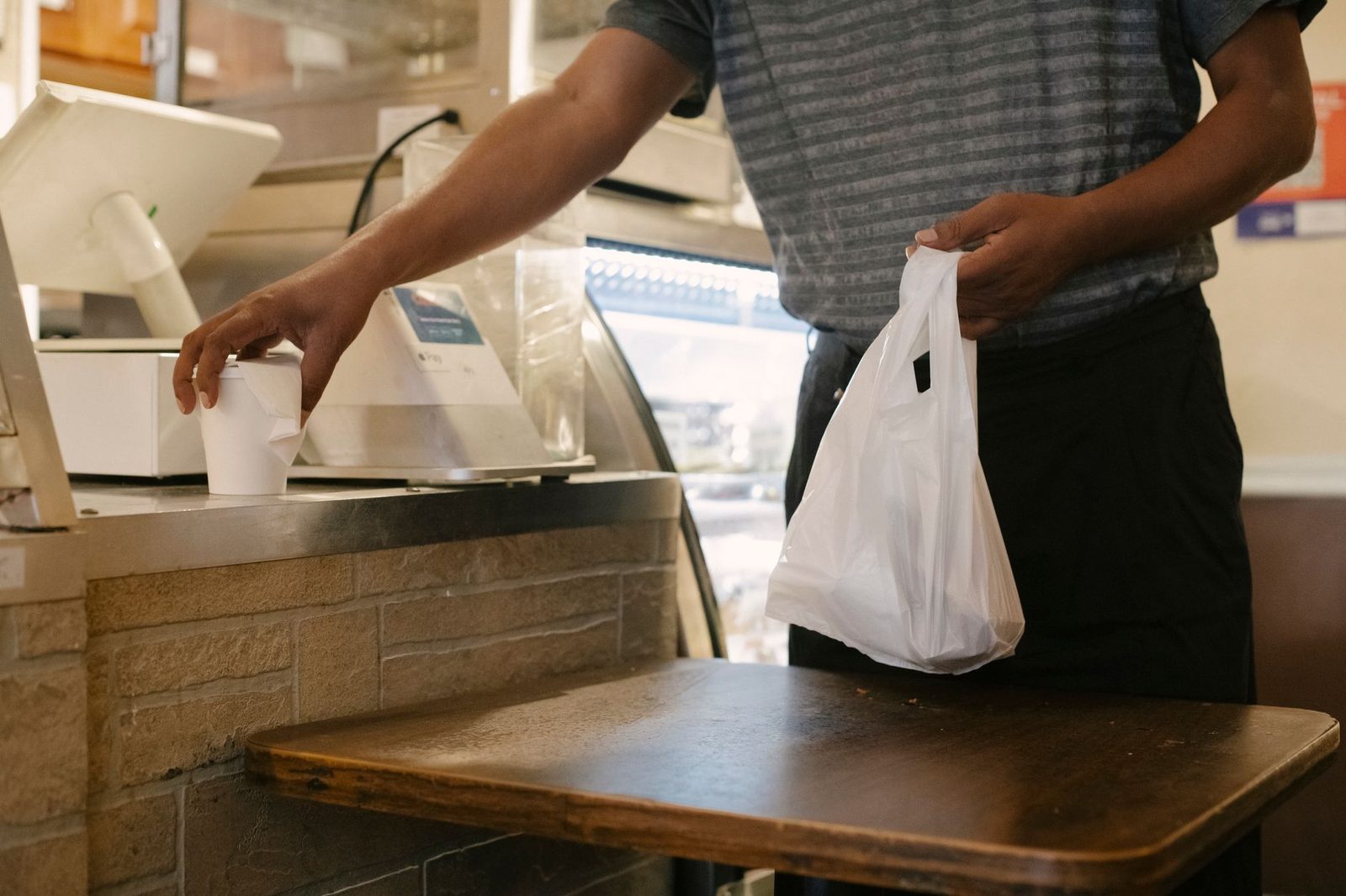 A man in casual clothes holds a plastic bag and a cup of hot drink at a cafe counter.