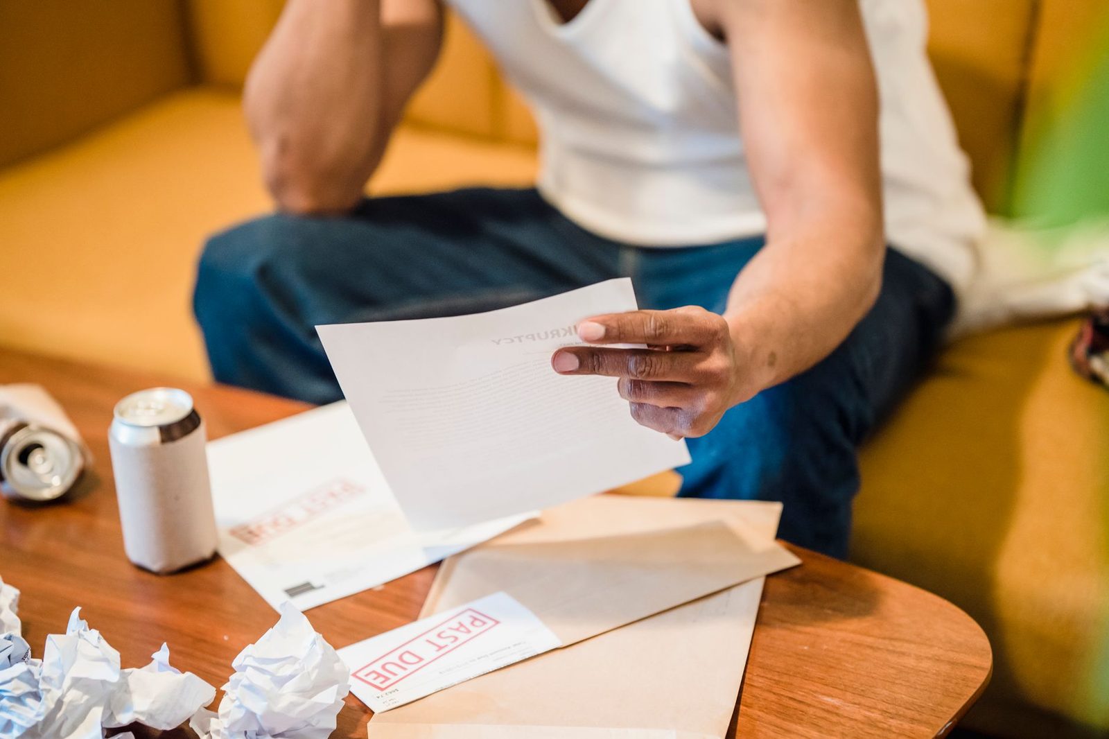 A man sits on a sofa, looking stressed as he reads through a stack of unpaid bills.