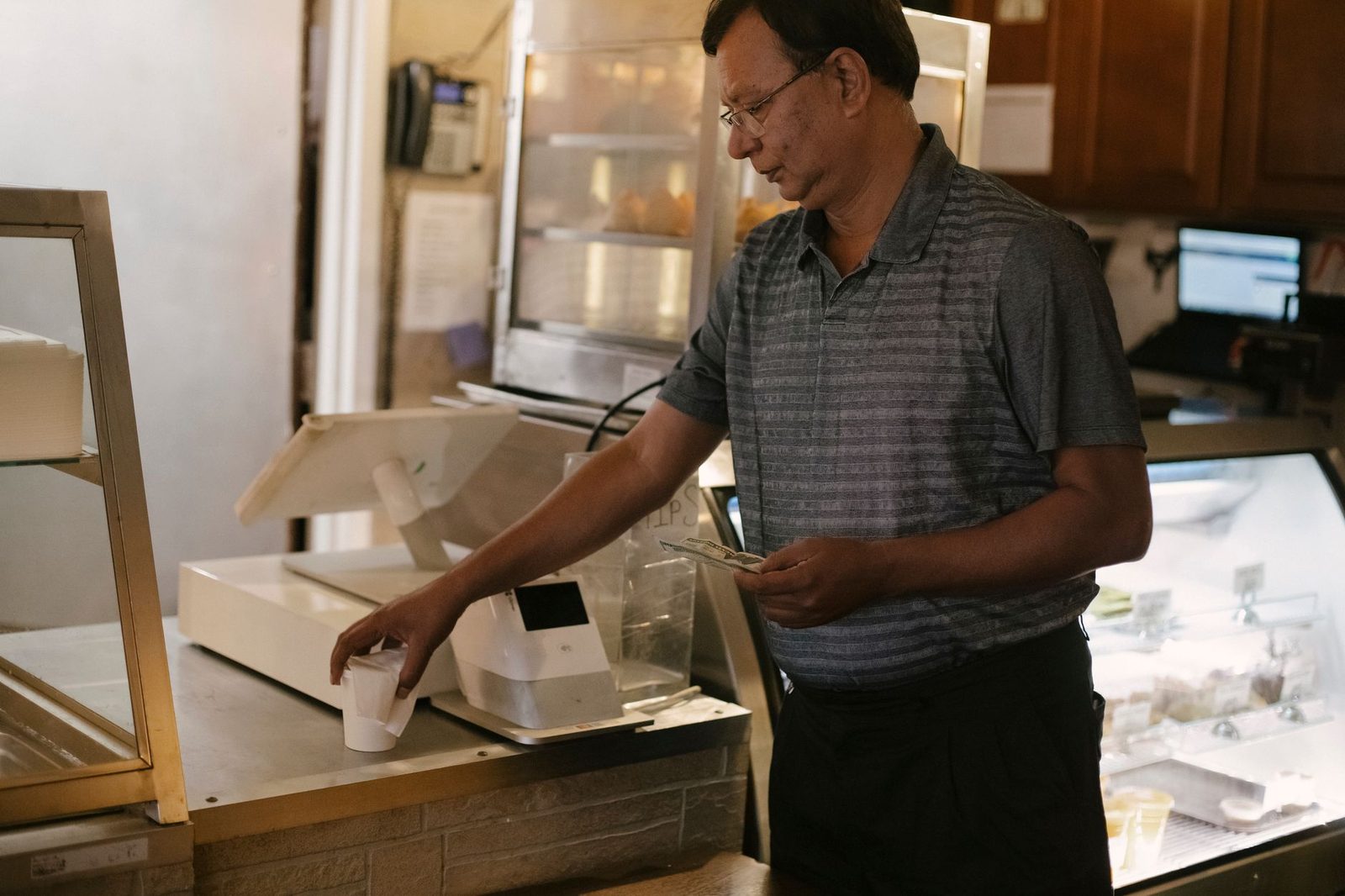 A man wearing eyeglasses picks up a cup of coffee beside a cash register and display case in a café.