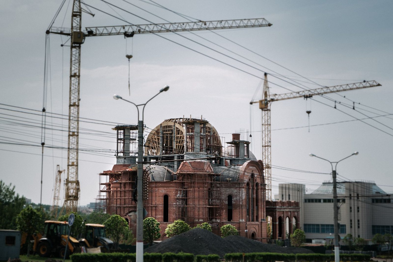 A red brick building under construction, with cranes rising above it against the sky.