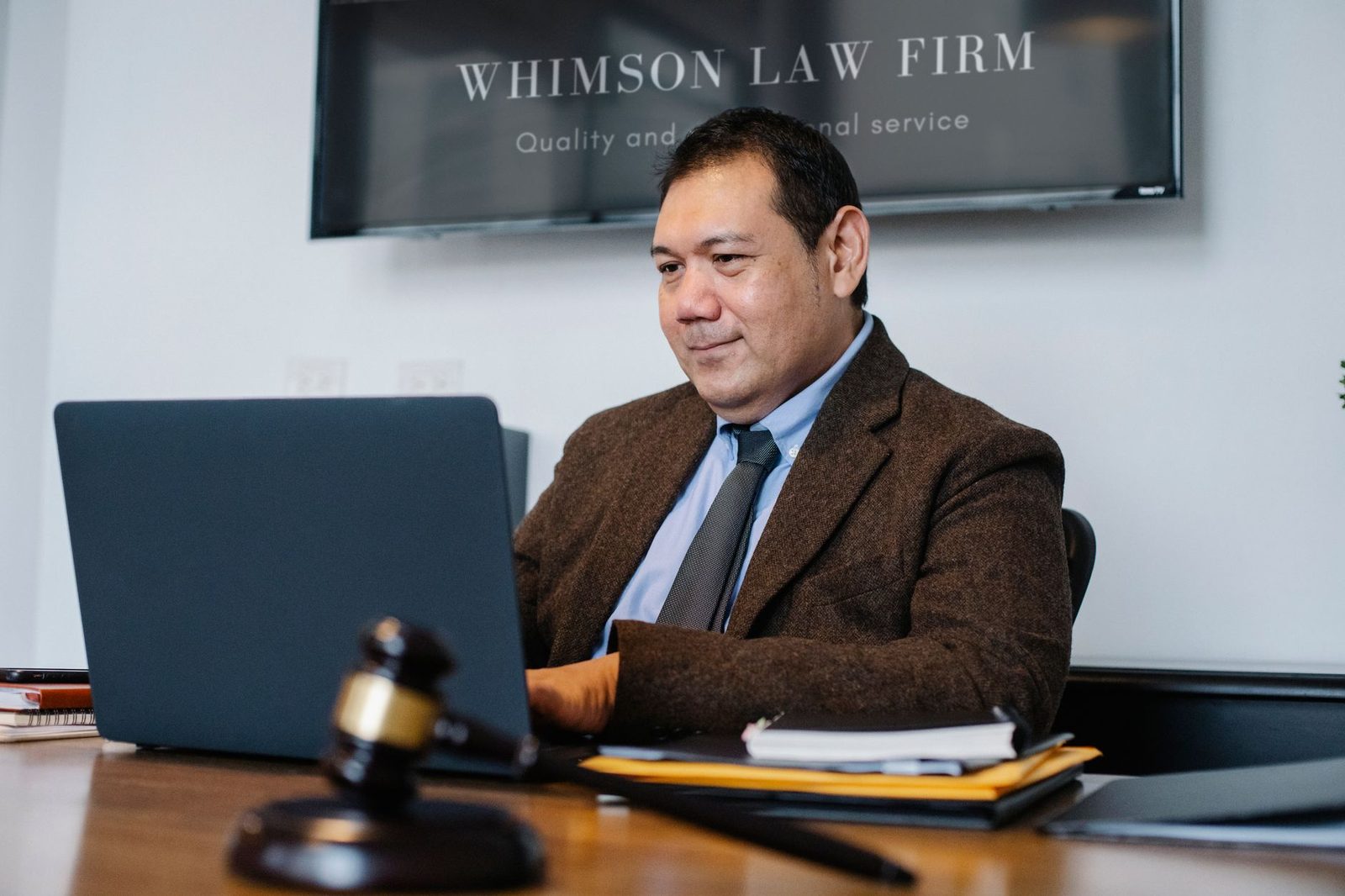A smiling middle-aged man works on a laptop at a desk beside a judge's gavel.