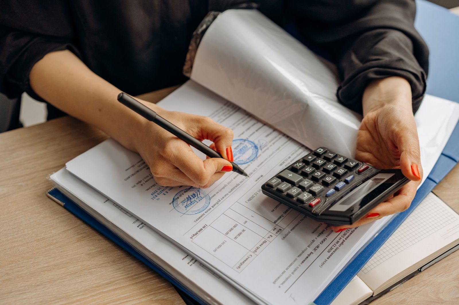An accountant uses a calculator beside a stack of documents while signing financial paperwork.