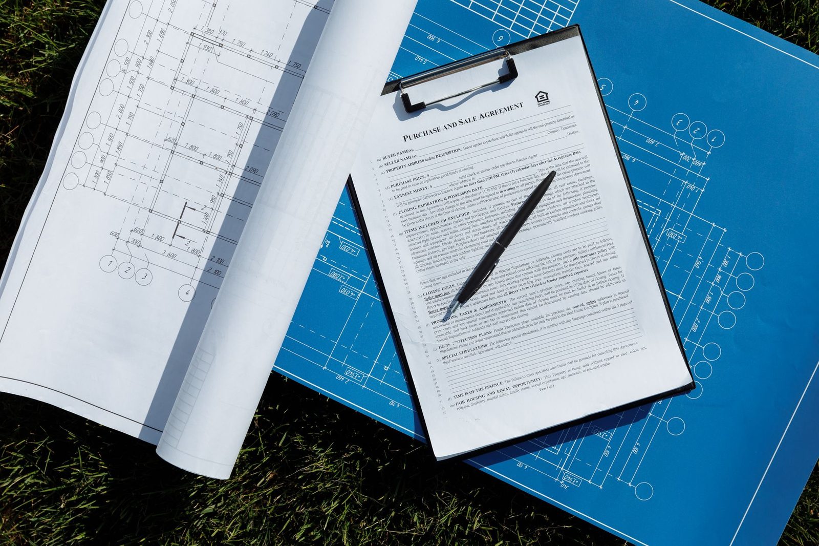 Blueprints and a signed contract with a pen lie spread out on the grass, viewed from above.