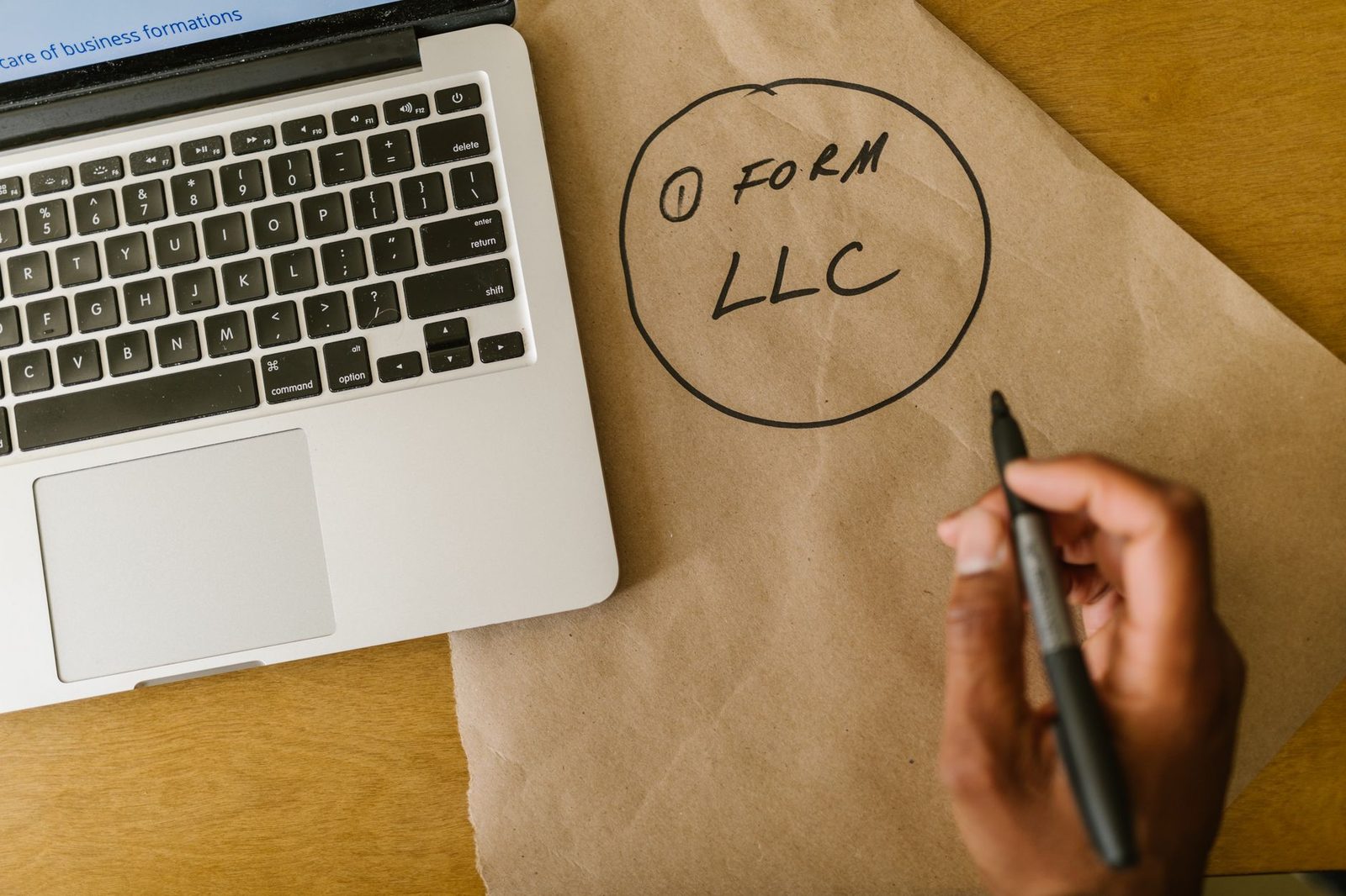 Primer plano de una mano escribiendo "Form LLC" en papel kraft junto a una laptop.