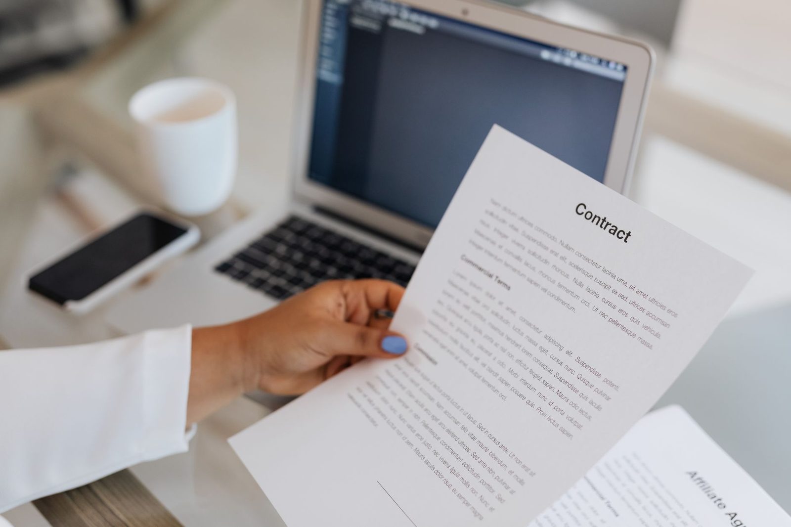 Close-up of a person holding a contract document beside a laptop during a work session.