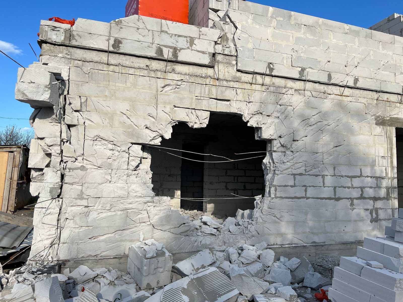 Concrete building wall with a large hole, rubble at its base, under a clear blue sky.