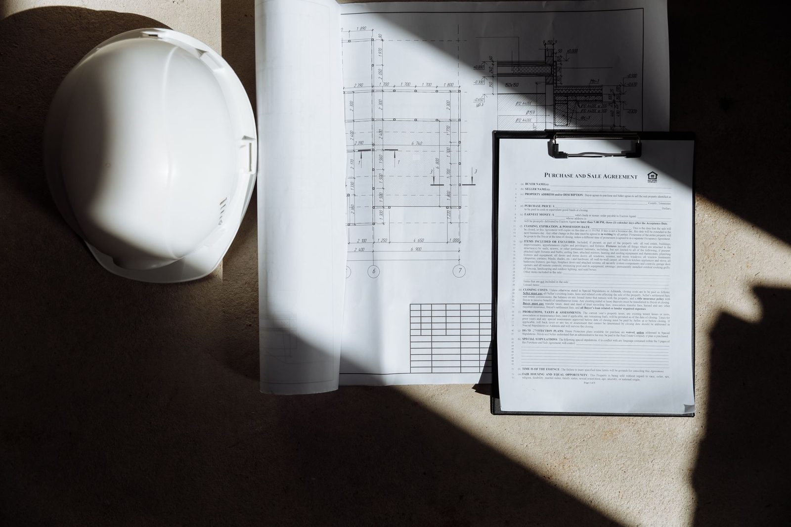 Construction blueprints, a hard hat, and a pen arranged on a desk in natural sunlight.