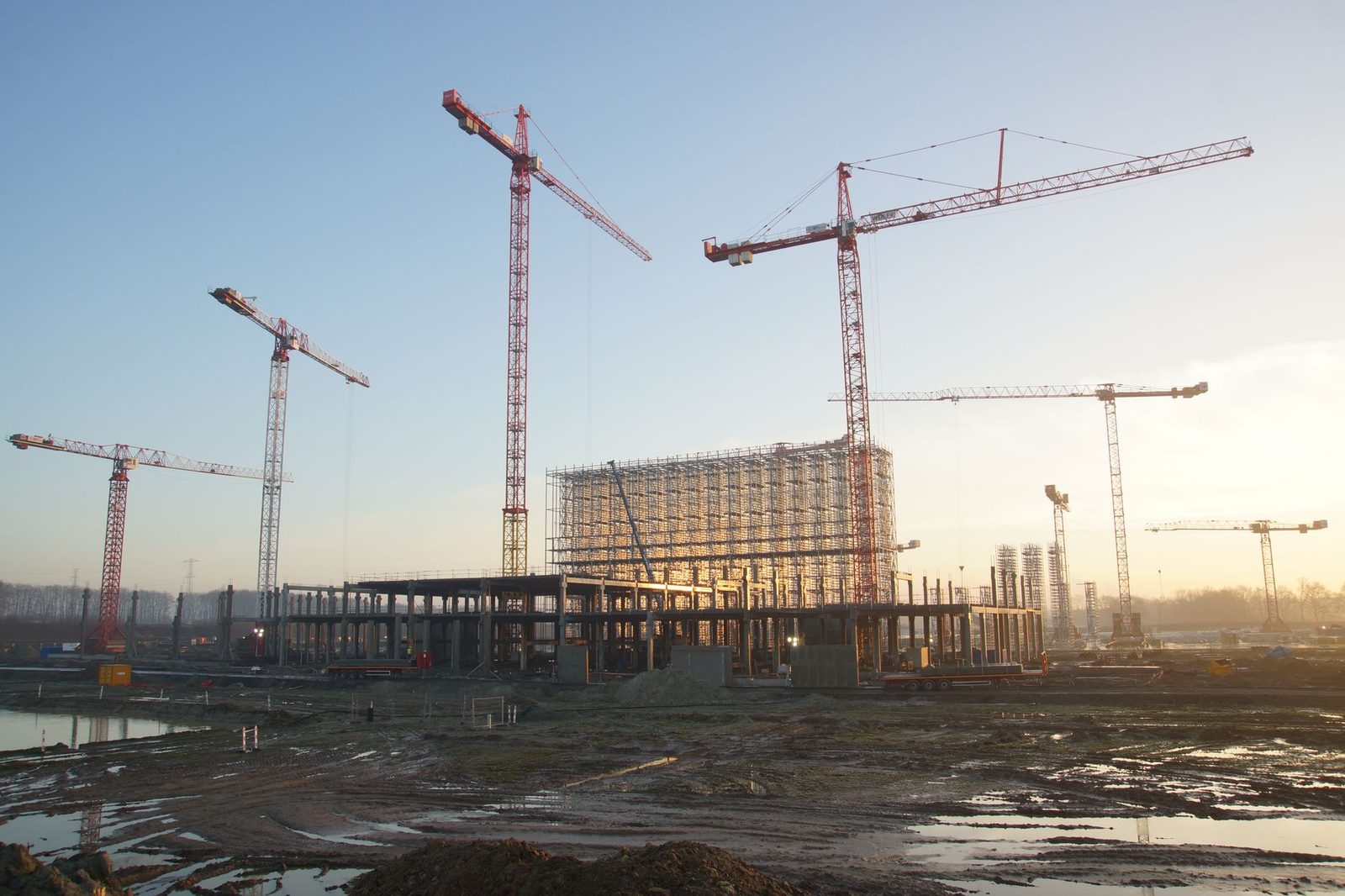 Construction site with cranes and steel framework silhouetted against a sunset sky.