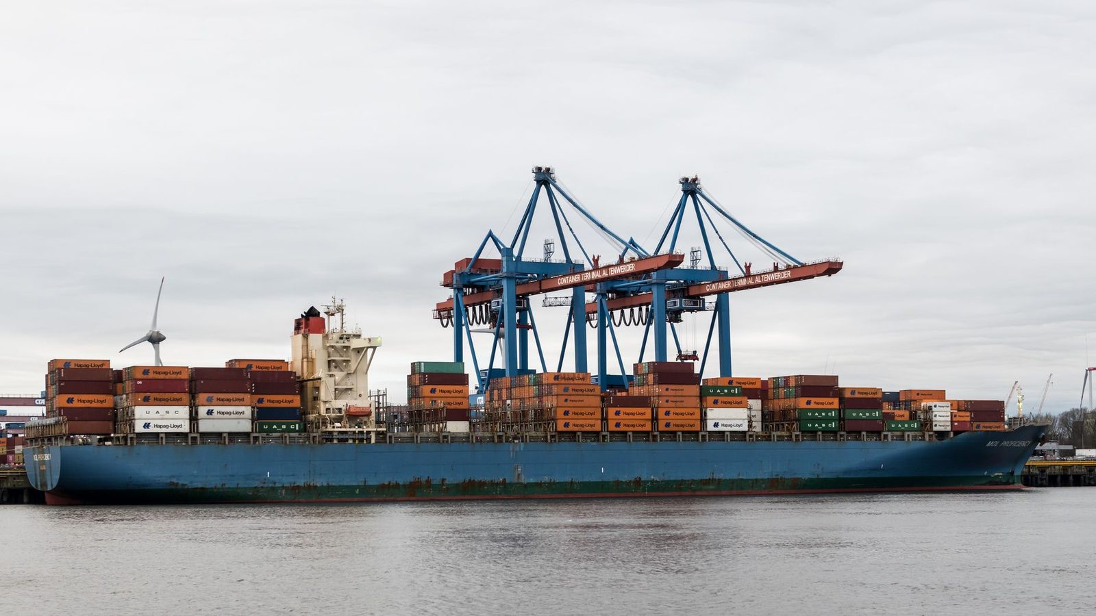 Container ship docked at a busy port, surrounded by tall cranes and stacked shipping containers.
