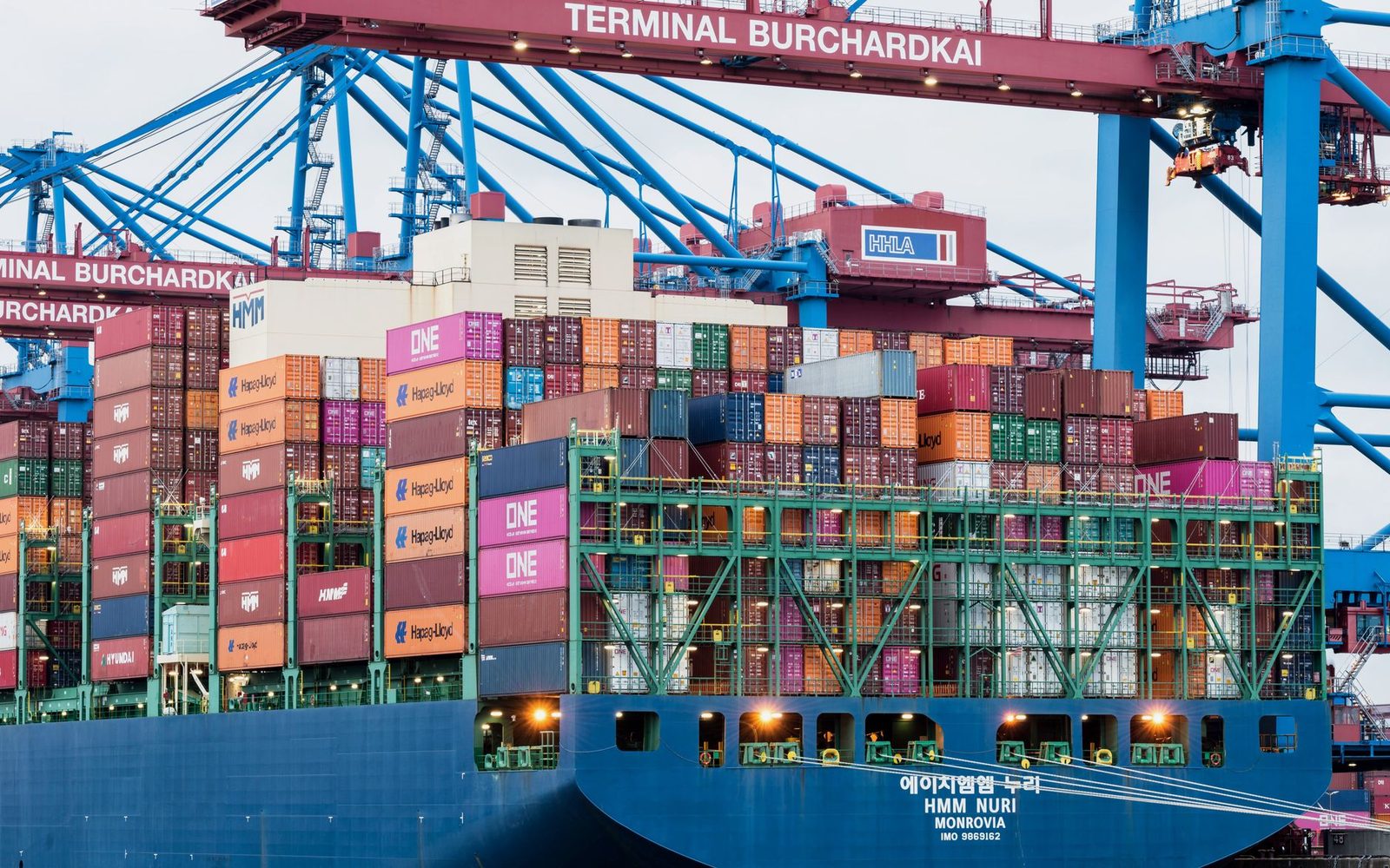 Container ship docked at a cargo terminal in Hamburg harbor, with stacked shipping containers and cranes.