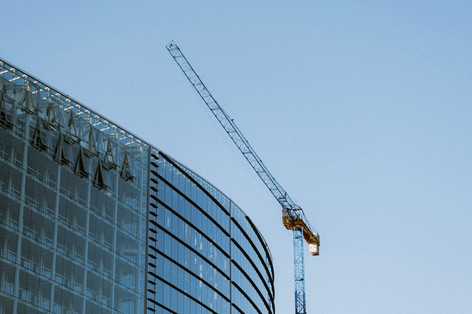A glass office building under construction with a crane against a clear blue sky.