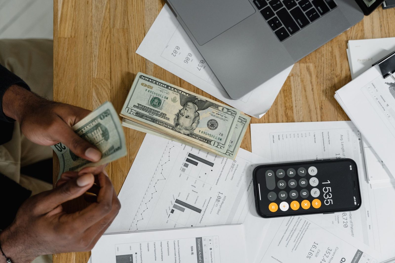 Hands counting cash beside a calculator, laptop, and printed charts on a desk.