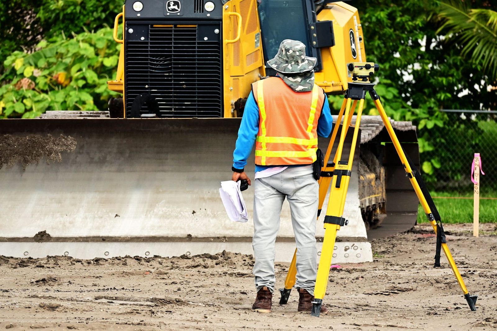 Surveyor with a tripod-mounted instrument observing bulldozer work at a Tampa Bay construction site.