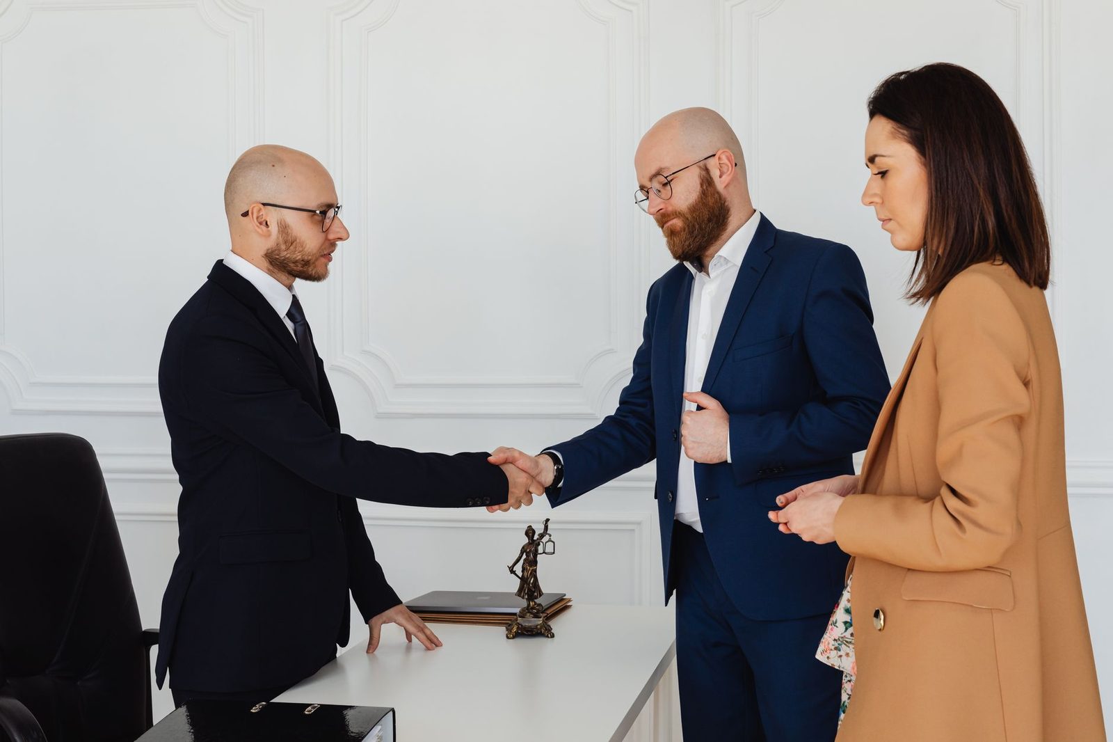 Three business professionals in formal attire shake hands across a table during an office meeting.
