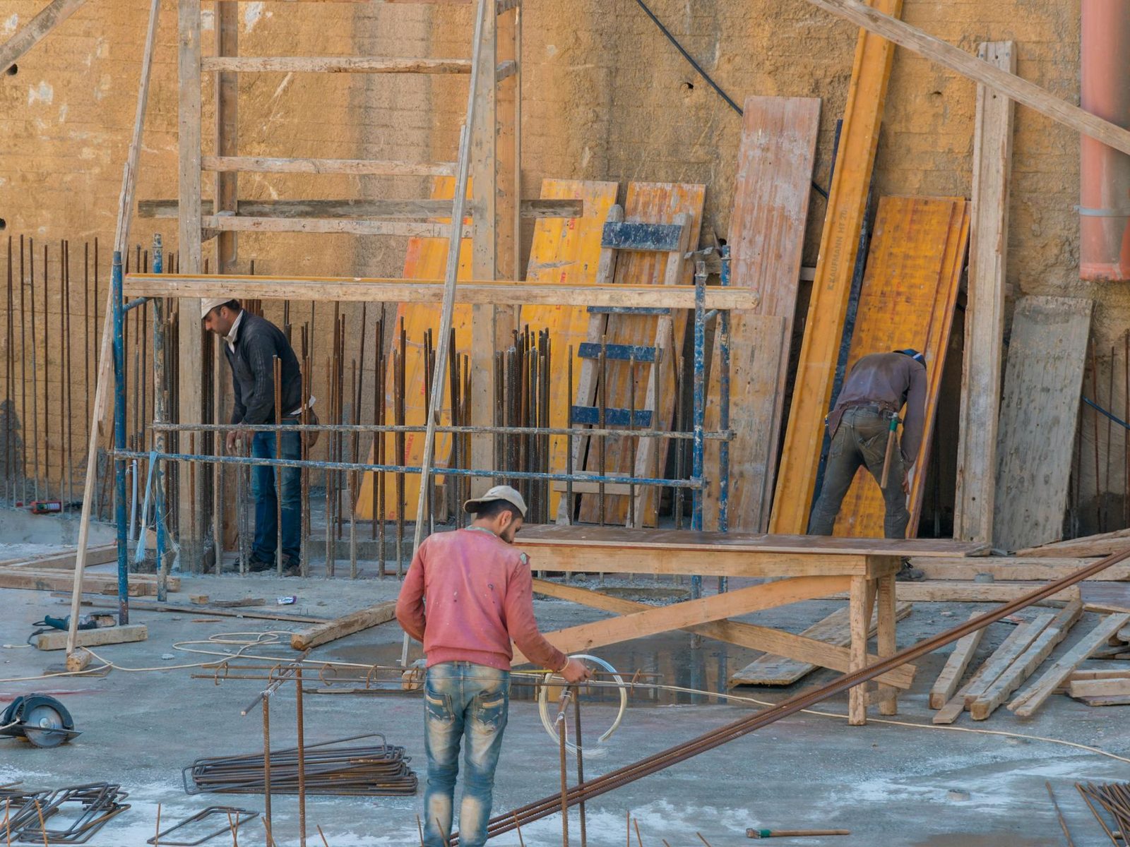 Three construction workers stand among wooden planks and metal rods on an outdoor building site.