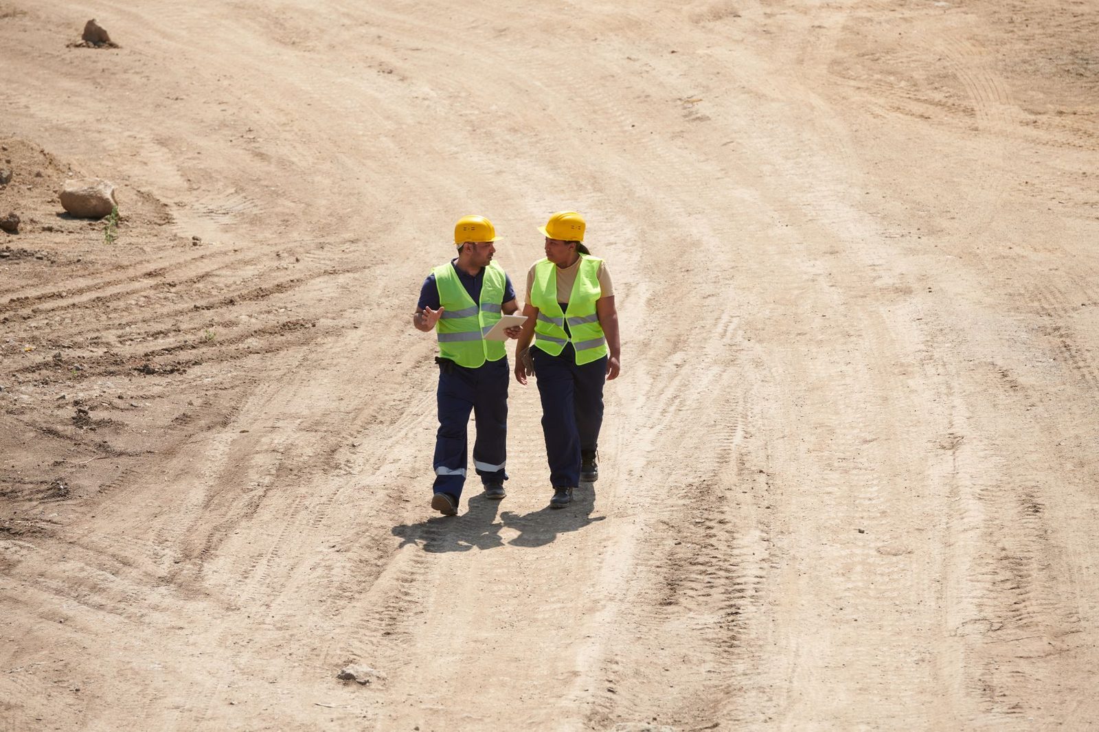 Two construction workers in safety vests and helmets discuss plans on a job site.