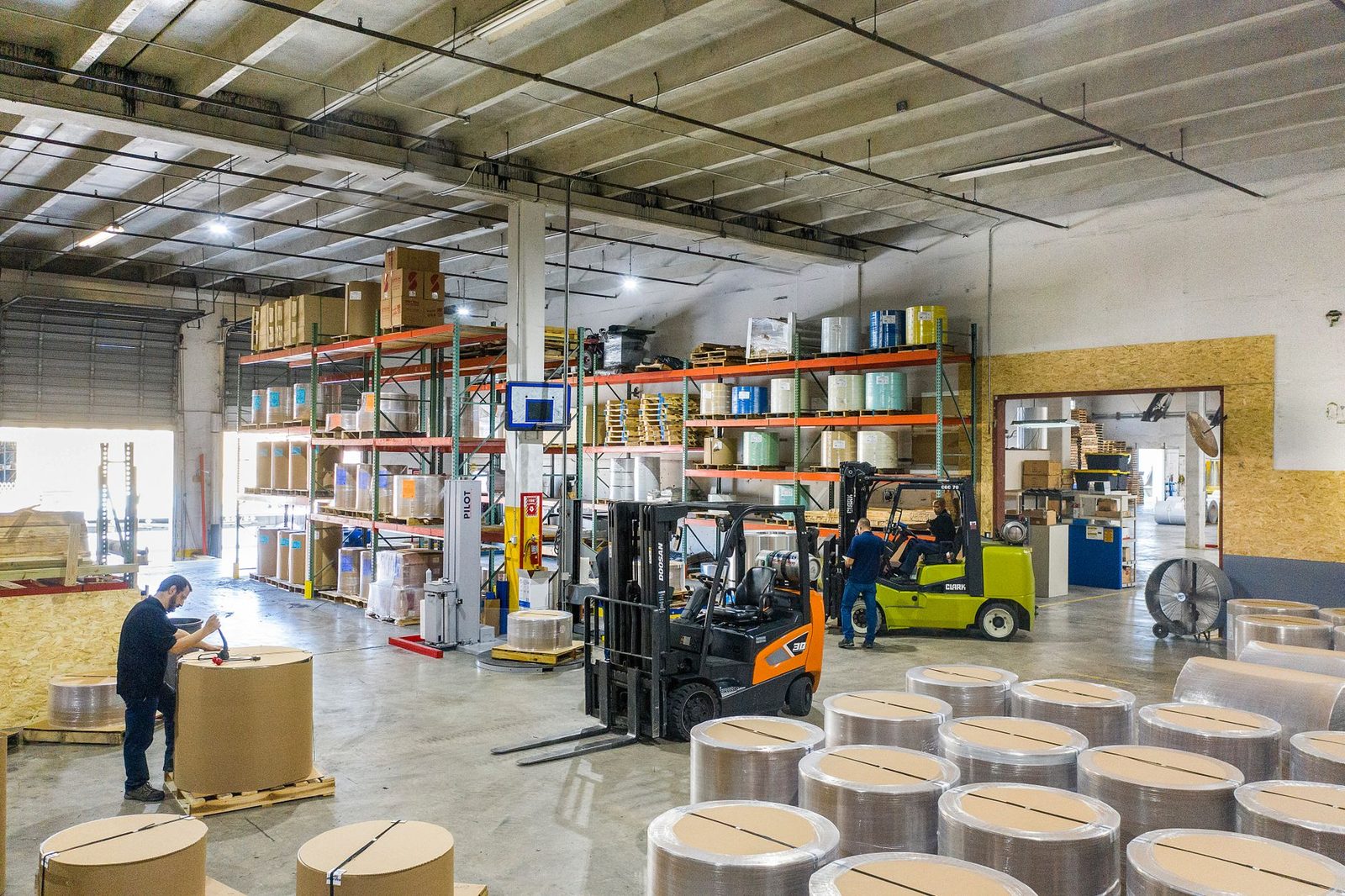 Workers and forklifts moving large material rolls through a busy warehouse interior.